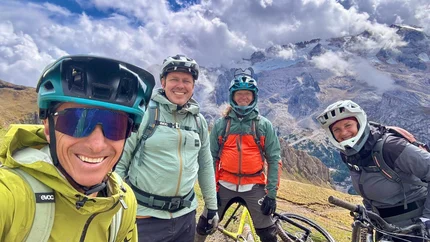 Four mountain bikers in helmets pose with their bikes against an alpine backdrop