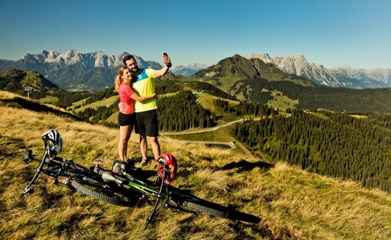 SUMMER IN THE MOUNTAINS Couple taking selfie on mountain with bikes and Alps in background