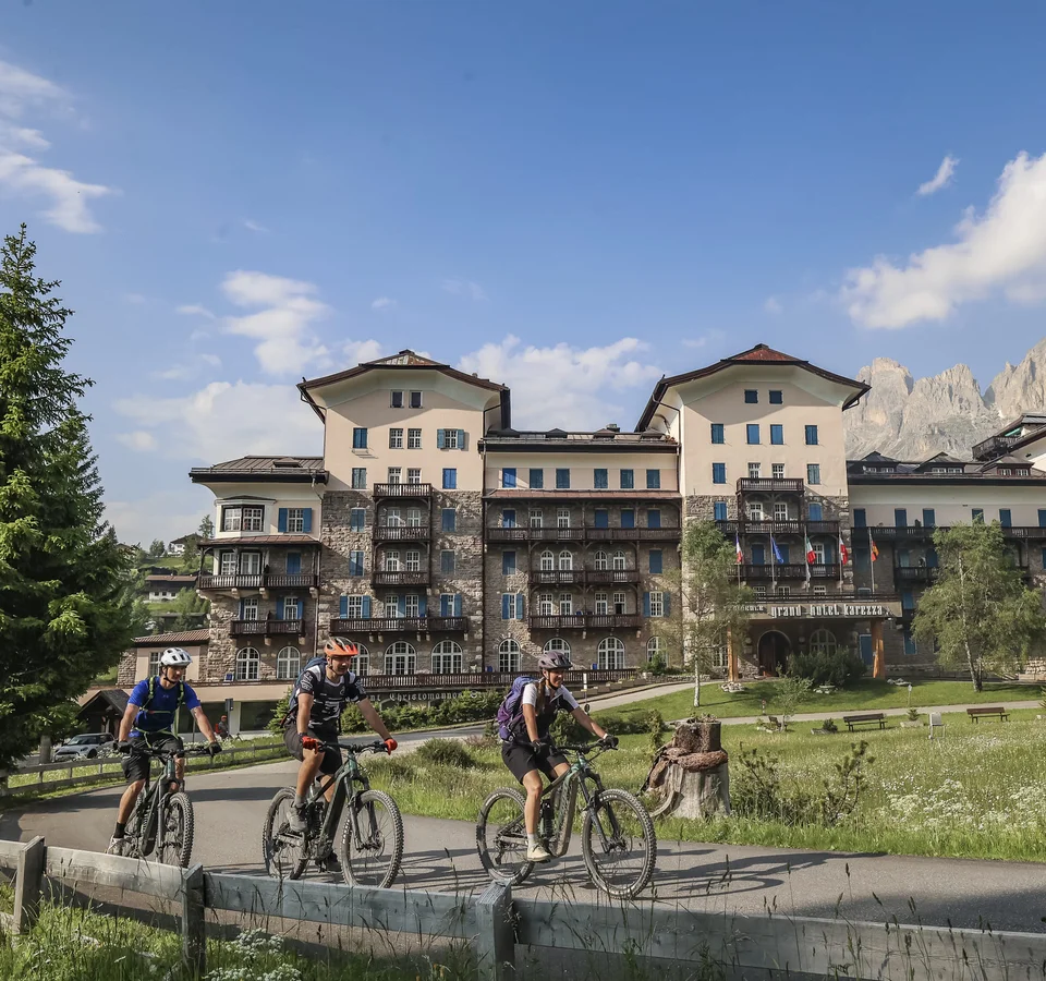 Genuss-(E-)Biken in den Dolomiten © Jens Vögele - Westsiders Drei Radfahrer vor einem großen Berghotel mit Bergen im Hintergrund