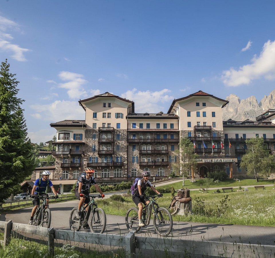 Drei Radfahrer vor einem großen Berghotel mit Bergen im Hintergrund
