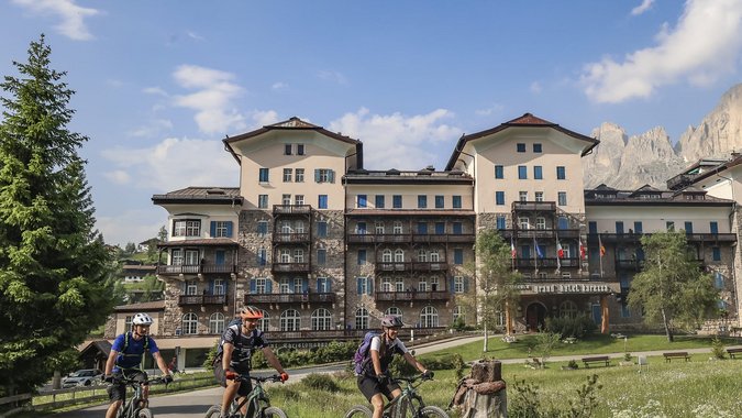 Drei Radfahrer vor einem großen Berghotel mit Bergen im Hintergrund