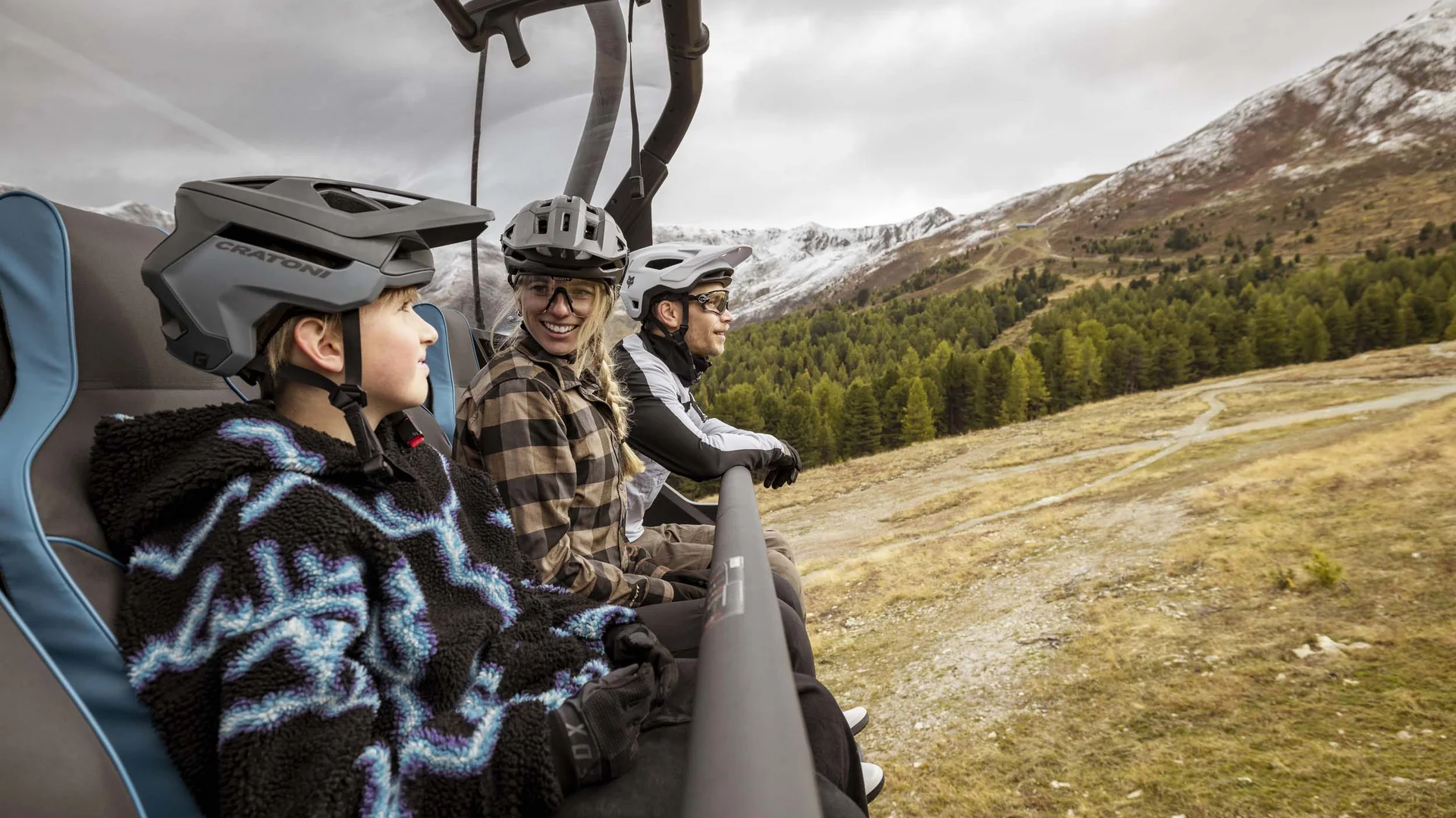 Drei Radfahrer mit Helmen in Sessellift über bergige Landschaft