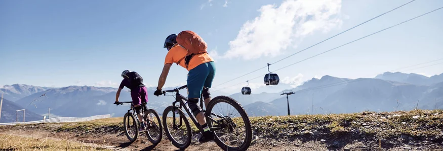 Two cyclists riding on a mountain trail under sunny sky