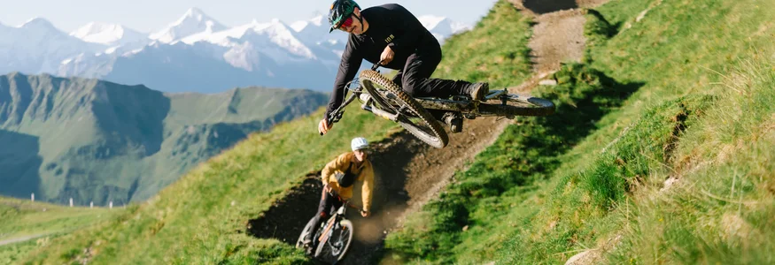 Am Westgipfel des Schattberg © saalbach.com/Moritz Ablinger Zwei Mountainbiker fahren einen Bergweg mit Gletscherbergen im Hintergrund