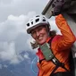 Smiling woman wearing helmet and orange jacket climbing a wooden structure in the mountains