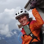Frau mit Helm und orange Jacke beim Klettern an Holzstruktur in den Bergen