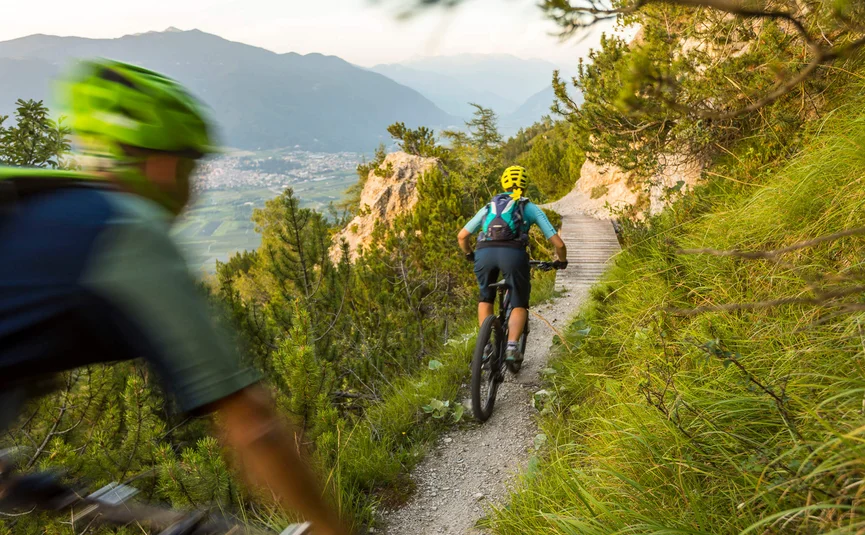 Two mountain bikers riding on a narrow mountain trail with green foliage