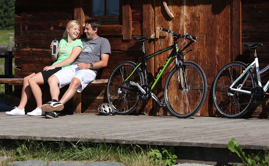 Paar sitzt lachend auf Bank vor Holzhütte mit Fahrrädern daneben