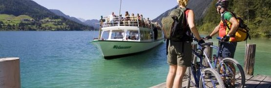 Twee fietsers op een steiger kijken naar een passagiersboot op een bergmeer