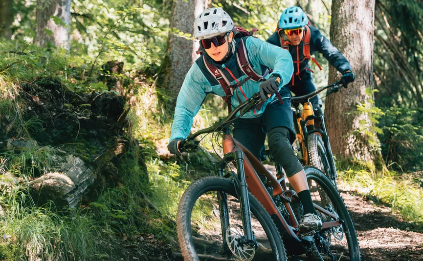 Zwei Mountainbiker fahren auf einem Waldweg über einen Baumstamm