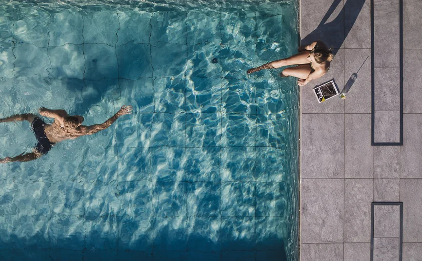 Man swimming in pool, woman sitting poolside with drink and magazine