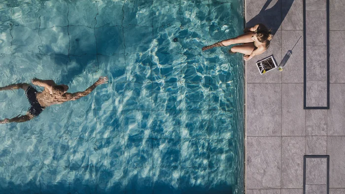 Mann schwimmt im Pool, Frau sitzt am Beckenrand mit Getränk und Zeitschrift