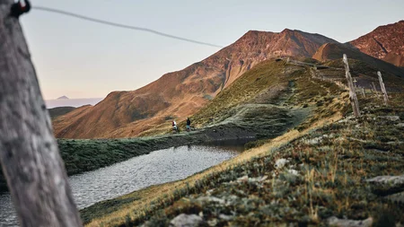Ready to ride: Das ist neu in Österreichs größter Bike-Region © Tobias Köhler Zwei Radfahrer an einem Bergsee mit Bergen im Hintergrund bei Sonnenuntergang