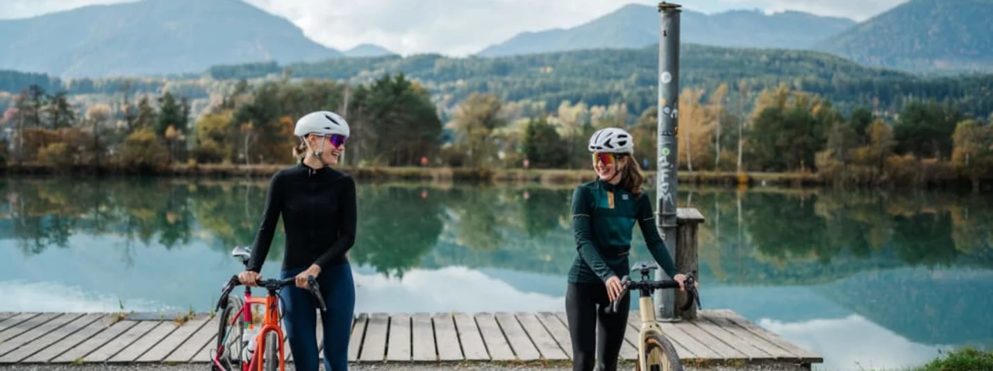 Two female cyclists with bikes by lake shore with mountains in background