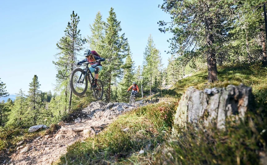 Mountainbiker springt auf felsigem Waldweg, ein anderer folgt hinten