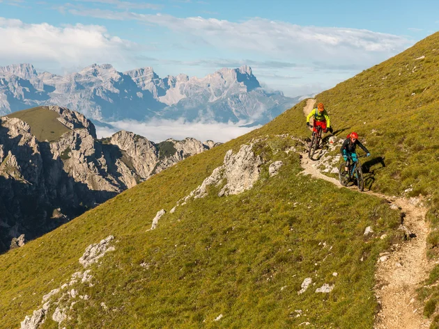 Zwei Mountainbiker auf einem schmalen Pfad in den Bergen unter blauem Himmel