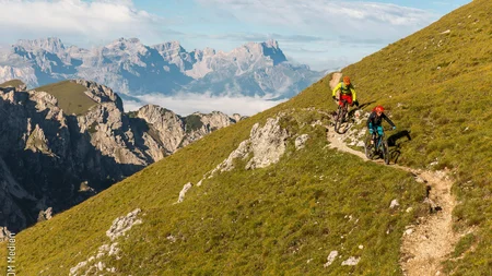 Zwei Mountainbiker auf einem schmalen Pfad in den Bergen unter blauem Himmel