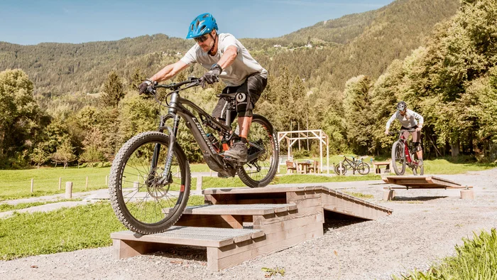 Mountainbiker fährt über eine Rampe im Gelände mit vielen Bäumen und Bergen