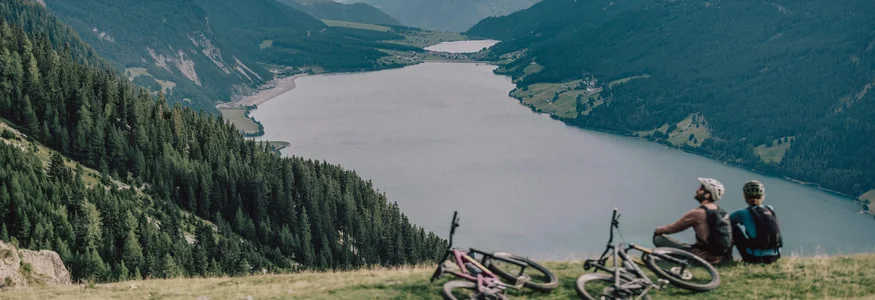 Zwei Radfahrer sitzen auf einem Hügel und blicken auf einen Bergsee und bewaldete Berge