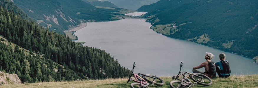 Green Days in Nauders-Reschensee © Rudi Whylidal Zwei Radfahrer sitzen auf einem Hügel und blicken auf einen Bergsee und bewaldete Berge