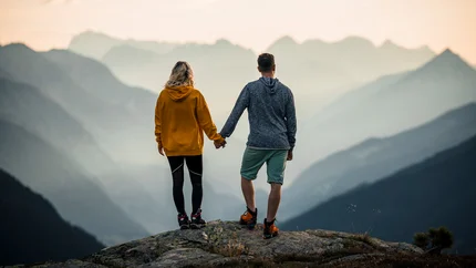 Paar hält Händchen auf Berggipfel mit Nebel und Bergpanorama im Hintergrund