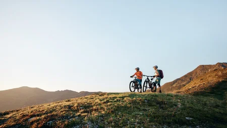 Unterwegs in Österreichs größter Bikeregion © Tobias Köhler Zwei Radfahrer mit Mountainbikes auf einem grasbewachsenen Hügel bei Sonnenuntergang