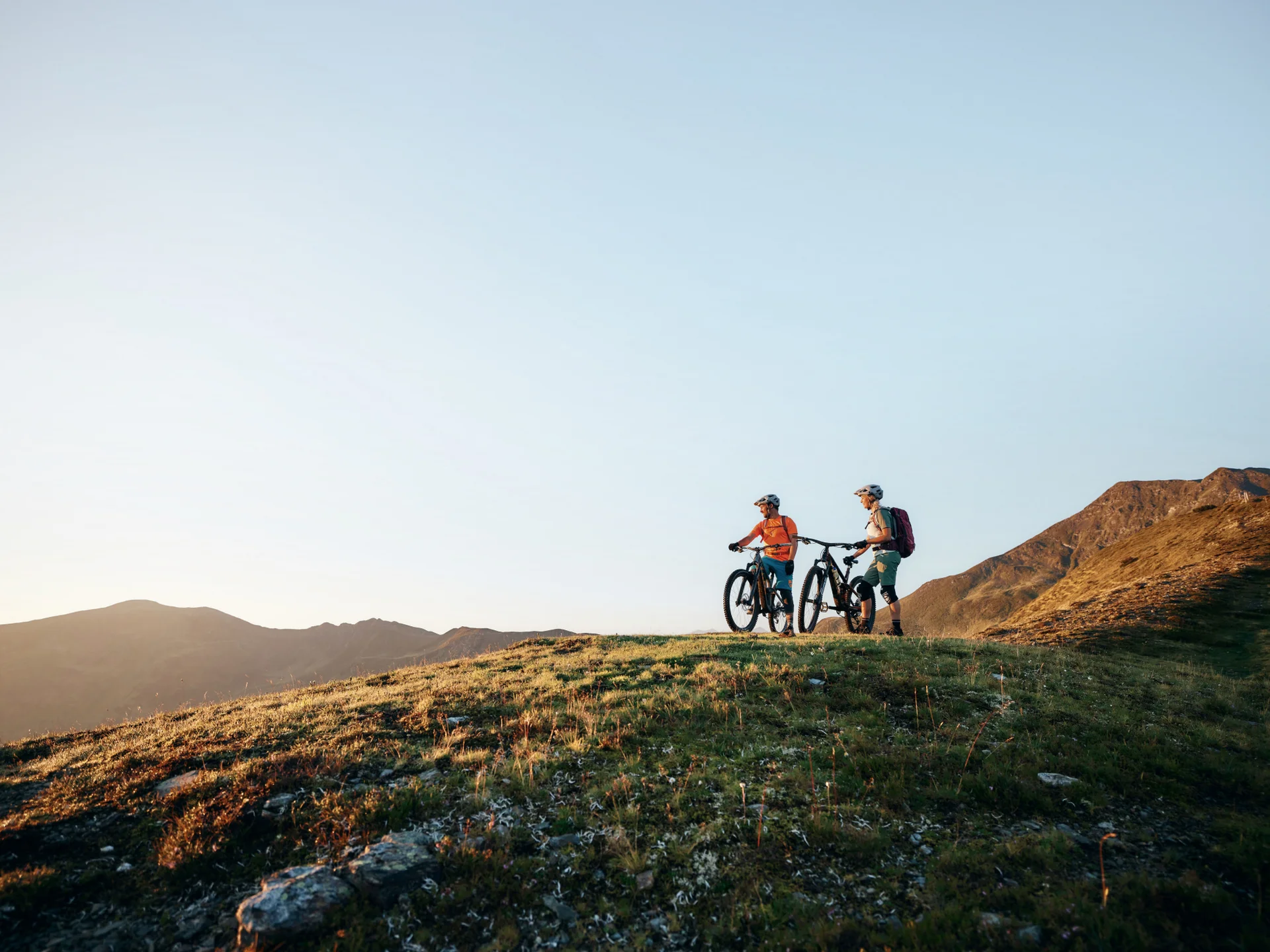Zwei Radfahrer mit Mountainbikes auf einem grasbewachsenen Hügel bei Sonnenuntergang