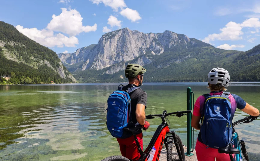Twee fietsers met helmen en rugzakken kijken uit over een bergmeer en bergen