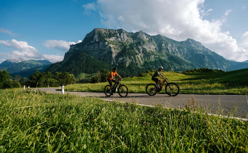 Zwei Radfahrer fahren auf einer Straße vor einem Bergpanorama bei Sonnenschein
