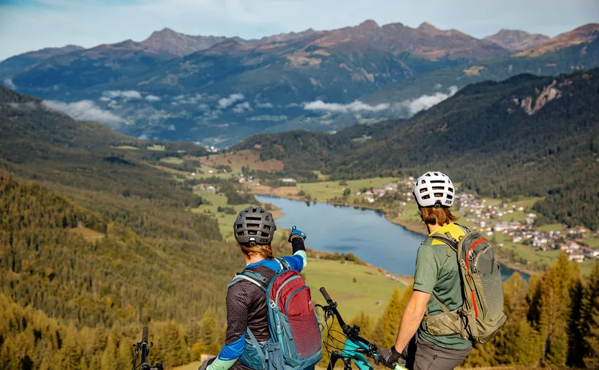 Zwei Mountainbiker mit Helmen blicken auf ein Tal mit See und Bergen