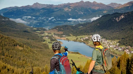 Zwei Mountainbiker mit Helmen blicken auf ein Tal mit See und Bergen