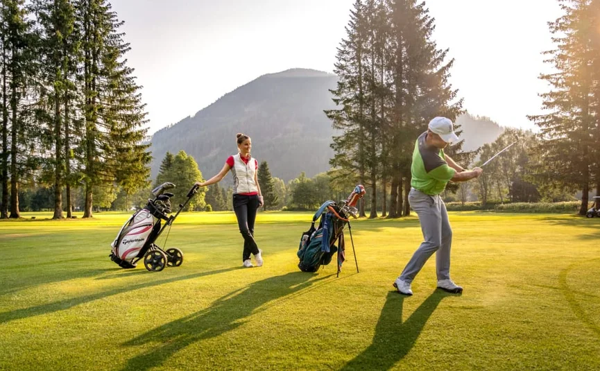 Mann schlägt Golfball, Frau zieht Golftrolley auf sonnigem Golfplatz