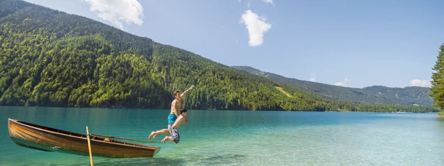 Twee jongens springen van een boot in een helder bergmeer met beboste heuvels