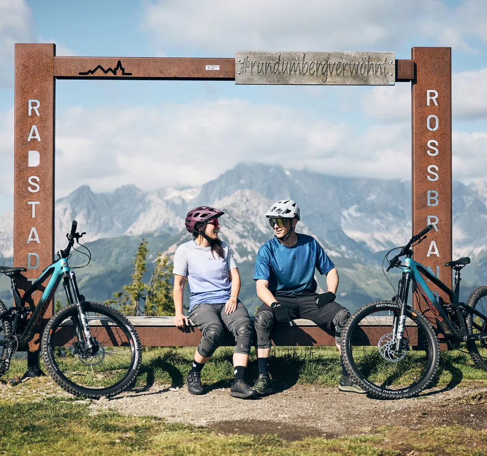 Bike-Region Flachau – von Radabenteuer bis Signature Ride © Tobias Köhler Zwei Radfahrer sitzen neben ihren Fahrrädern an einem Bergblick-Rahmen in Radstadt Rossbrand.