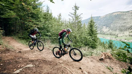 Twee mountainbikers op een bospaadje met uitzicht op bergen en een meer