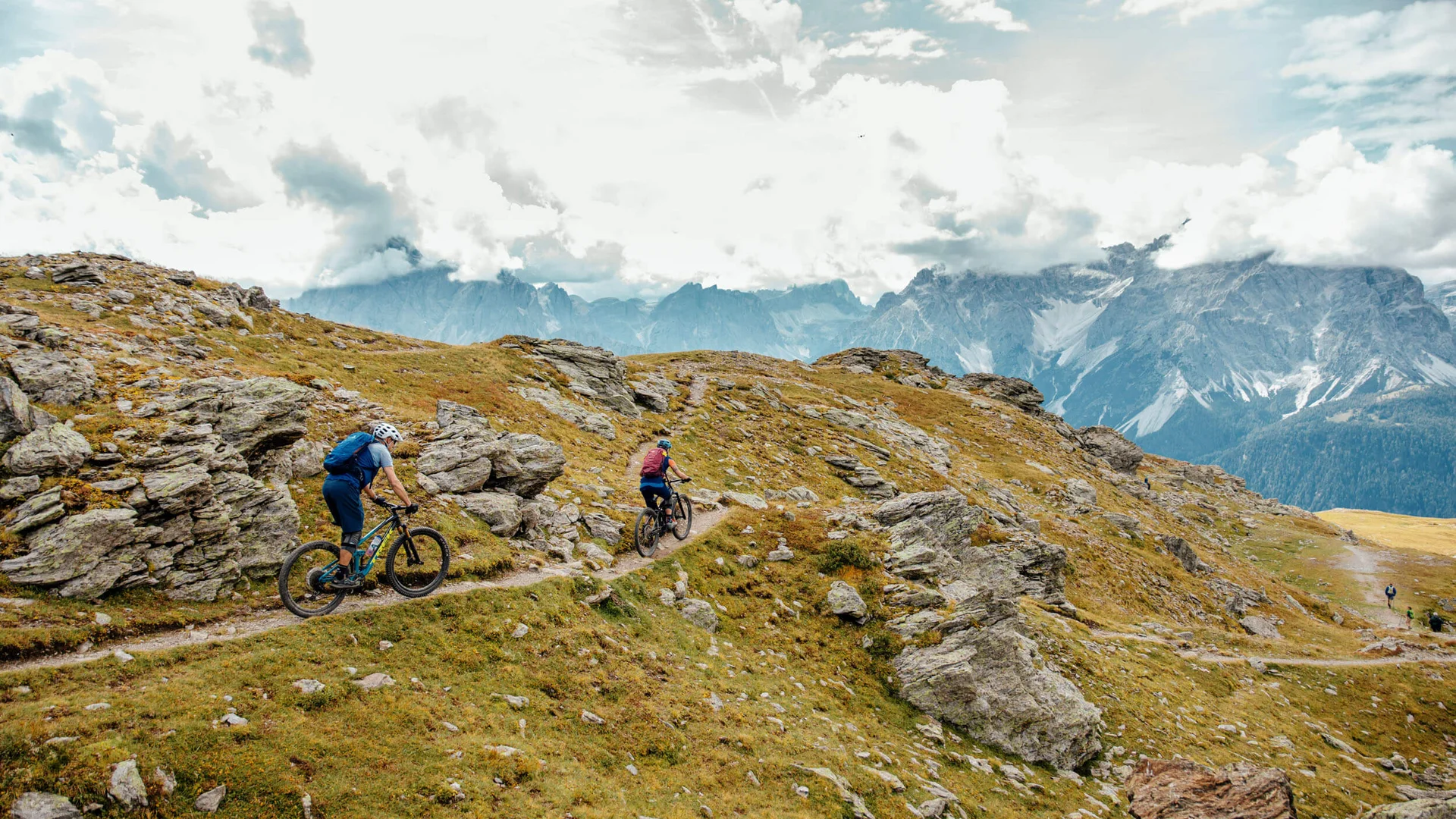 Two mountain bikers riding on a rocky mountain trail under cloudy sky