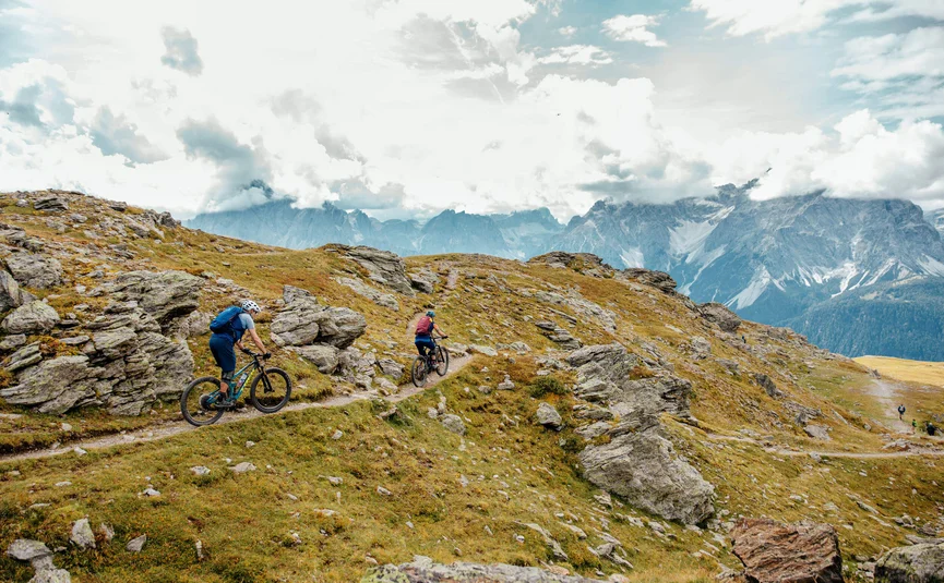 Two mountain bikers riding on a rocky mountain trail under cloudy sky