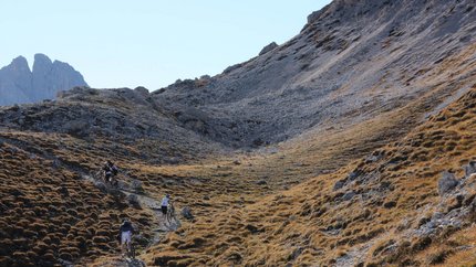 Drie personen duwen fietsen omhoog op een rotsachtig bergpad bij zonnig weer