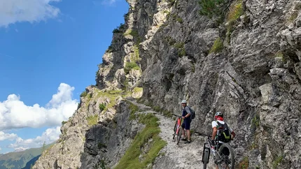 Zwei Radfahrer schieben ihre Mountainbikes auf einem Bergpfad entlang einer steilen Felswand