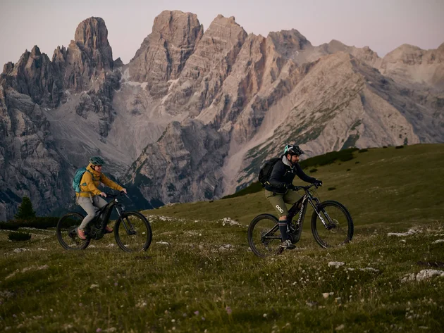 Was steckt hinter dem Umweltengagement von Bikehotels? © David Rasche Zwei Mountainbiker fahren auf einer Wiese vor hohen Bergen