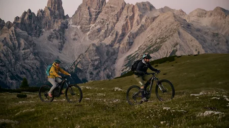 Was steckt hinter dem Umweltengagement von Bikehotels? © David Rasche Zwei Mountainbiker fahren auf einer Wiese vor hohen Bergen