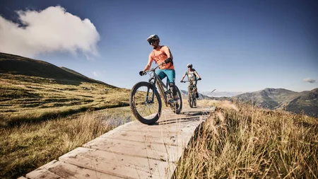Unterwegs in Österreichs größter Bikeregion © Tobias Köhler Zwei Mountainbiker fahren auf einem Holzsteg in den Bergen bei klarem Himmel