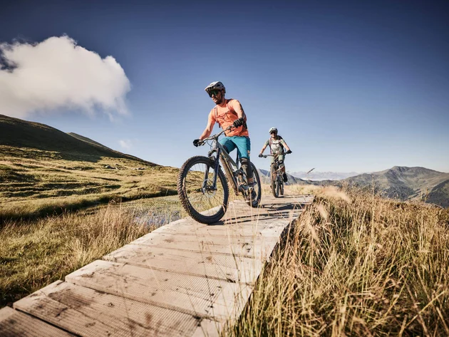 Unterwegs in Österreichs größter Bikeregion © Tobias Köhler Zwei Mountainbiker fahren auf einem Holzsteg in den Bergen bei klarem Himmel