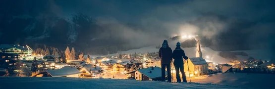 Verschneites Bergdorf bei Nacht mit zwei Menschen und beleuchteter Kirche