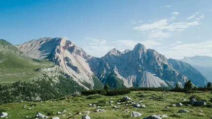 Grüne Wiese mit Felsen vor steilen Bergen unter blauem Himmel