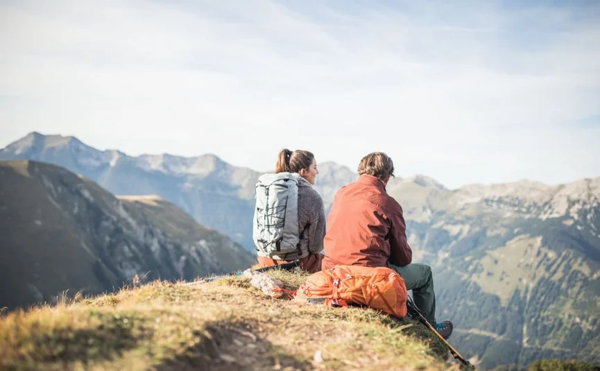 Twee wandelaars zitten op een berg met rugzakken uitkijkend over de Alpen