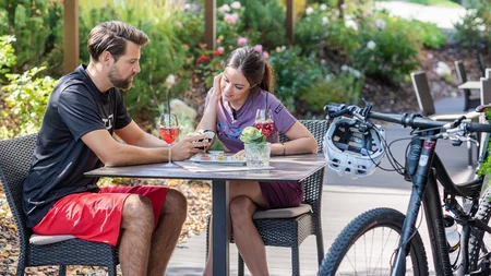 Pärchen auf Terrasse mit Fahrrädern, trinkt Getränke und schaut auf Smartphone