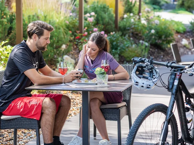 Pärchen auf Terrasse mit Fahrrädern, trinkt Getränke und schaut auf Smartphone
