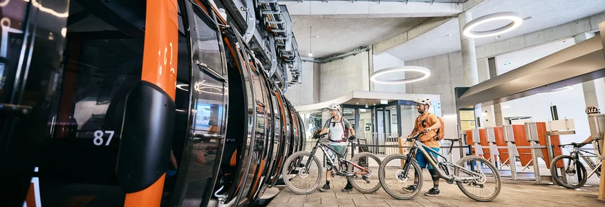Zwei Radfahrer mit Helmen warten in einer modernen Seilbahnhaltestelle