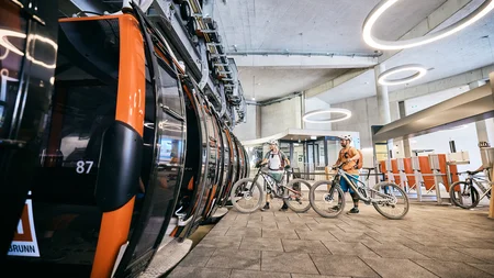 Zwei Radfahrer mit Helmen warten in einer modernen Seilbahnhaltestelle